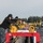 170113-N-GS192-181 A group of U.S. Navy personnel in digital camouflage uniforms are operating a large torpedo loading or handling apparatus on what appears to be a pier or deck facility. The equipment consists of a long rail-mounted system with yellow mechanical drive mechanisms and a large cylindrical tube (likely a torpedo or torpedo tube component) positioned on the red-framed cradle. The setting appears to be a naval installation, possibly in the Pacific Northwest based on the conifer trees visible in the background.