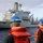 USS Lake Champlain (CG 57) Replenishment-at-Sea Three naval personnel wearing blue hard hats and orange life vests are observing a large gray replenishment vessel from the deck of another ship at sea. The scene appears to depict a replenishment at sea (RAS) or underway replenishment (UNREP) operation, with the supply ship positioned alongside. The weather is overcast with moderate sea conditions.