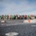 170123-N-MJ135-152 The image shows a large group of personnel assembled on what appears to be a naval aircraft carrier flight deck. In the foreground, an arresting wire cable and deck hardware with an orange triangle marker are in sharp focus, while the crowd and a carrier island structure are visible in the background. Personnel are wearing various colored jerseys/vests typical of carrier flight deck crew roles.