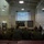 HMH-464 Marines attend Road to War Brief during Exercise Frigid Condor A military briefing or training session is taking place in what appears to be a military facility or gymnasium. Personnel in military uniforms are seated facing a projection screen displaying a 'Questions?' slide marked 'Overall Classification: UNCLASSIFIED'. Two individuals stand at the front of the room conducting the presentation.