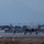 Lightning II strikes Iwakuni, F-35B arrives Two stealth fighter jets, consistent with F-35 aircraft, are taxiing on a military airfield runway at dusk or dawn in low-light conditions. The scene shows an active flight line with runway lighting illuminated and ground personnel visible. Several military hangars and support structures are visible in the background.