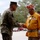 Navajo code talker visits VMU-2 A U.S. Marine Corps officer in camouflage utility uniform shakes hands with an elderly veteran wearing a red cap inscribed 'Navajo Code Talkers' and a gold/orange shirt adorned with military ribbons and Native American jewelry. The meeting appears to be a formal or ceremonial greeting, likely at a military installation. A second Marine in uniform is visible in the background.