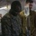 2d Medical Bn Field Exercise A U.S. Marine in MARPAT camouflage uniform with the name tape 'MARITIM' is standing near an IV drip stand inside what appears to be a military field medical tent. A second individual wearing an olive/khaki uniform and a stethoscope is visible in the background, suggesting a medical training or treatment scenario. The setting appears to be a forward operating base or field hospital environment.