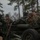 Artillery Training School Three military personnel in camouflage uniforms and combat helmets are operating or standing near a large artillery piece, likely an M777 howitzer or similar towed artillery system. The setting appears to be an outdoor training area with pine trees in the background under an overcast sky. The soldiers appear to be conducting artillery training or preparation exercises.