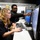 CyberThon 2017 Two individuals are working together at a computer workstation in what appears to be a technology lab or classroom. A young woman with curly blonde hair is seated at the desk operating a mouse, while a young man wearing glasses and a black hoodie stands behind her, pointing at one of the monitors. The setting includes multiple computer setups with monitors, keyboards, and headsets.