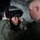 From bolts to bombs Two individuals in olive/green military flight suits are shown in what appears to be a military aircraft interior. One person is assisting the other in fitting or adjusting a black flight helmet with a visor. The interaction suggests a pre-flight equipment check or training scenario.