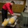 170118-N-BL637-046 A sailor or ordnance technician wearing a red long-sleeve shirt and dark pants is working on what appears to be practice/training bombs (inert ordnance, indicated by yellow stripe markings) secured on a cart. The setting appears to be the hangar deck or weapons elevator area of a U.S. Navy aircraft carrier, as evidenced by the 'KEEP OFF THIS ELEVATOR WHEN IN OPERATION' warning sign and yellow-black hazard striping. The individual appears to be inspecting or securing the ordnance with chains or straps.