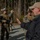 Defeat the Device A civilian instructor or advisor in a black jacket and tan baseball cap appears to be briefing or instructing a group of military personnel in woodland camouflage uniforms. The soldiers are equipped with body armor, helmets, and rifles, suggesting a training exercise in a forested outdoor environment. The setting appears to be a military training area with a gravel surface and pine trees in the background.