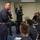 170123-N-GZ228-017 A senior military official in dark blue coveralls addresses a group of U.S. Navy sailors seated in a classroom setting. The sailors are wearing the Navy Working Uniform (NWU) digital camouflage pattern. The setting appears to be a briefing or training session aboard a naval vessel or installation.
