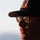 USS Cheyenne hosts bussines leaders A close-up profile shot of a man wearing a dark baseball cap with an embroidered crest/logo and rectangular-framed glasses. The image is taken during golden hour lighting, casting warm directional light across the right side of his face. The background shows a pale blue sky with a faint horizon line, suggesting an outdoor elevated or coastal setting.