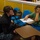 NPASE COMREL A person in U.S. Navy digital camouflage (NWU) uniform is crouching beside a young child seated at a small school desk. The adult appears to be assisting or tutoring the child, who is looking down at a worksheet or printed document on the desk. The setting appears to be a classroom or educational facility with yellow walls and blue student chairs.