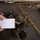 Marines train while aboard USS Mesa Verde Military personnel, appearing to be U.S. Marines based on their distinctive MARPAT camouflage uniforms and gear, are gathered on what appears to be a ship's deck or hangar bay floor. One individual is holding a spiral-bound notebook with handwritten entries in a grid/checklist format, suggesting an administrative or accountability task is being conducted. Personnel are equipped with full combat gear including helmets, body armor, and rifles.