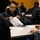 170119-N-OY799-039 A group of military personnel in Navy Working Uniform (NWU) digital camouflage are seated at tables in what appears to be a briefing or administrative processing event. A female sailor in the foreground, with a name tape partially visible reading 'SAUND...', is opening or handling an envelope marked 'FOR OFFICIAL USE ONLY'. Multiple sailors are engaged with paperwork in the background.