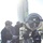 Expeditionary ordnance loading exercise U.S. Navy personnel in blue digital camouflage uniforms and hard hats are handling what appears to be a torpedo or large cylindrical ordnance on a loading cradle/skid on the deck of a submarine. The crew members are guiding the weapon using ropes and direct handling, with one sailor visible wearing a name tape reading 'JACKSON'. A second person in the background appears to be monitoring the operation with a clipboard.