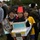 Service Members compete in Ginowan Traffic Safety Relay Race Three children pose together at what appears to be an outdoor school event or parade. The central child wears a handmade cardboard vehicle costume with the number '55' and '110' displayed on it, along with a red bucket on their head. They are holding a sign with Japanese text indicating '55 - 葛飾小学校 6年4組' (Katsushika Elementary School, 6th Grade Class 4).