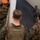 Marines train while aboard USS Mesa Verde The image shows multiple military personnel in camouflage uniforms and tactical gear inside what appears to be a ship's well deck or cargo bay. The central figure, identified by a name tape as 'JOHNSON,' is viewed from behind wearing a plate carrier vest over MARPAT (Marine pattern) camouflage, suggesting U.S. Marine Corps affiliation. The personnel appear to be staging or moving through the enclosed space during a training exercise or operational deployment.