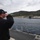 170123-N-JI086-220 A person wearing a dark navy jacket and cap with an American flag patch stands on the deck of a naval vessel, shading their eyes to observe a harbor facility in the distance. The harbor contains what appear to be one or two submarines moored at a pier, set against a hillside with Mediterranean-style vegetation. The scene suggests a naval port visit or approach to a submarine base.