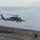 170118-N-NV908-043 A military helicopter, consistent with an MH-60 Seahawk, is hovering or landing on the flight deck of a naval vessel at sea. Two deck crew members are visible on the flight deck, with one appearing to signal the helicopter. The ocean and partly cloudy sky form the background.
