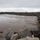 Camp Pendleton Flooding 2017 A swollen, turbulent river with muddy brown water is shown adjacent to a road bridge or causeway with metal guardrails. The water level appears elevated, suggesting recent heavy rainfall or flooding conditions. Vehicles are parked or stopped on the roadway, and large riprap rocks line the embankment in the foreground.