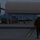 Lightning II strikes Iwakuni, F-35B arrives A military personnel member wearing camouflage uniform and a flight deck helmet walks across a tarmac toward aircraft shelters at dusk or dawn. In the background, what appears to be an F-35 stealth fighter jet is visible parked under or near a large open-sided aircraft shelter. The scene appears to be at a military air base, likely during low-light conditions.
