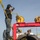 170113-N-GS192-136 U.S. Navy personnel in digital camouflage uniforms are operating mechanical equipment on what appears to be a naval vessel or pier facility. A sailor in the foreground is operating a yellow hand-crank winch mechanism mounted on a red steel frame, while a large cylindrical tube with internal rifling or spiral grooves is prominently visible in the foreground. The setting appears to be a waterfront naval installation in cold weather conditions.