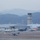 Lightning II strikes Iwakuni, F-35B arrives A military airbase is depicted at dusk or dawn with hazy atmospheric conditions, showing multiple aircraft on the tarmac and a prominent air traffic control tower in the background. A single-engine stealth-type jet aircraft (consistent with an F-35 profile) is visible in the foreground on the runway, while several maritime patrol and transport aircraft are parked near hangars. Mountains are visible in the background, suggesting a location in a mountainous coastal region, consistent with a Japanese military installation.