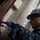 170118-N-BR087-047 A U.S. Navy sailor wearing a white hard hat marked '74', safety glasses, and blue nitrile gloves is working on a red pipe or hose fitting aboard a naval vessel. The individual is dressed in Navy Working Uniform (NWU) digital camouflage pattern. The setting appears to be an interior compartment or passageway of a ship, with riveted white-painted steel bulkheads visible in the background.
