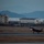 Lightning II strikes Iwakuni, F-35B arrives A stealth fighter jet, consistent with an F-35 variant, is captured during takeoff or landing roll on a military airfield runway at dusk or dawn. The base appears to be coastal, with water and mountainous terrain visible in the background. Military support buildings and infrastructure are visible behind the runway.