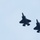 Lightning II strikes Iwakuni, F-35B arrives Two dark-colored fifth-generation stealth fighter jets are flying in close formation against a pale blue-gray overcast sky. The aircraft are photographed from below at an angle, showing their ventral surfaces. Both jets appear to be flying in the same direction in a loose echelon formation.