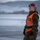 Boots in the Snow A female military police (MP) soldier stands at attention on what appears to be a tarmac or airfield in winter conditions. She is wearing camouflage fatigues, a red beret, and a high-visibility orange vest marked 'MP', indicating a military police traffic or security control role. The background shows a snow-covered landscape with mountains, suggesting a Nordic or northern European location.