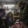 2d Medical Bn Field Exercise Military personnel in camouflage uniforms are conducting what appears to be a combat casualty care training exercise inside a field medical tent. Two individuals are actively working on a simulated casualty on a treatment table, with visible simulated blood and bandaging materials. Several other personnel observe the procedure in the background.