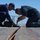 170122-N-PP996-024 Two military personnel in navy blue uniforms are kneeling on what appears to be a ship's deck, performing maintenance or inspection work on a track or rail system embedded in the deck surface. One individual is using a brush or tool to apply a substance or clean the track area, while the other observes closely. A green tool bag is visible nearby, and a safety net is visible in the background.