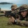 31st Marine Expeditionary Unit completes MEUEX Two military personnel in digital camouflage uniforms and boonie hats are lying prone on grass in a firing position, aiming rifles toward an unseen target. Both individuals and their weapons are covered in sand, suggesting they recently emerged from or trained near the beach visible in the background. The setting appears to be a coastal training exercise.