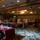 Fifth Annual Baby Expo An indoor vendor fair or craft market is taking place in a large hotel ballroom or convention hall. Multiple vendor tables covered with red/magenta tablecloths are arranged throughout the space, with attendees browsing displayed merchandise. The venue features ornate crystal chandeliers, recessed lighting, and a decorative patterned carpet.
