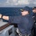 170121-N-KB426-048 Two naval personnel are standing on the bridge wing or exterior deck of a military vessel at sea. The foreground figure, wearing a cap with gold officer insignia and sunglasses, is gesturing outward with an extended arm, apparently pointing or directing attention to something in the distance. A second sailor stands behind him with arms crossed, observing.