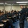 170123-N-GZ228-006 A senior U.S. Navy officer in Navy Working Uniform (NWU Type I, blue digital camouflage) addresses a large group of sailors seated at classroom-style desks. The setting appears to be a briefing or training room aboard a naval vessel or installation. A second uniformed sailor stands in the background near the instructor's desk.