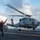 170123-N-BL637-071 Two naval aviation personnel are directing a military helicopter on the flight deck of an aircraft carrier at sea. One crew member wearing a green vest labeled 'LSE' and another in a brown jacket with a red helmet marked '7H' are signaling the helicopter during landing or takeoff operations. The rotor blades are visibly blurred due to rotation, indicating the helicopter is in active operation.