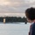USS Cheyenne hosts bussines leaders A submarine is navigating on the surface of a calm waterway, observed by a person in the foreground whose back is to the camera. The submarine's sail (conning tower) is visible with personnel standing on the deck, suggesting a port arrival or departure. The setting appears to be a tropical naval base, with lush green trees and low-lying structures visible on the shoreline in the background.
