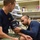 170123-N-TI693-011 Two individuals in navy blue shirts are in a medical or first aid room, where one person is taking the other's blood pressure using a manual cuff and stethoscope. A vital signs monitor is visible in the background displaying readings. The setting appears to be a clinical or emergency services environment based on the equipment and supplies visible on the shelves.