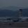 Lightning II strikes Iwakuni, F-35B arrives A military/joint-use airfield photographed at dusk or dawn with low ambient light. A fighter-type aircraft is visible on or near the runway in the foreground, with multiple parked aircraft, hangars, and an air traffic control tower visible in the background. Mountainous terrain forms the backdrop, suggesting an East Asian location.