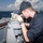 170121-N-KB426-053 A naval servicemember in dark blue uniform and cap is using a pelorus or bearing circle mounted on the bridge wing of a military vessel at sea. The individual is looking through the sighting vane of the navigation instrument, likely taking a bearing or fixing the ship's position. The setting is an open ocean environment with partly cloudy skies visible in the background.