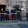 170118-N-SK327-039 A group of six naval personnel dressed in dark blue uniforms and caps are working together on the deck of a naval vessel, handling a large thick mooring rope or line. They are arranged in a line, each gripping the rope and appearing to haul or position it across the deck. The setting is clearly a military ship, with gray metal superstructure, pipes, electrical boxes, and an open hatch visible in the background.