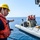 170116-N-LR795-036 A naval or coast guard operation is depicted aboard a military vessel at sea. A sailor wearing a yellow hard hat and orange life vest appears to be directing or communicating with a group of personnel seated on what appears to be a rigid inflatable boat (RIB). The personnel on the boat are wearing tactical gear, helmets, and orange life vests, suggesting a boarding team or special operations deployment.