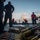 170123-N-VN584-049 The image shows military personnel on the deck of a naval vessel, with linked ammunition belts prominently displayed in the foreground. Several crew members wearing red shirts and dark coveralls are visible, some kneeling and working near what appears to be a deck-mounted weapon system. The scene appears to depict a weapons drill or live-fire exercise at sea during early morning or dusk conditions.