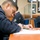 USS Lake Champlain (CG 57) CPO Advancement Exam The image shows multiple individuals seated at wooden desks, writing on paper with pencils in what appears to be a classroom or testing environment. All visible individuals are wearing matching dark navy blue uniforms or sweatshirts, suggesting an institutional or military/law enforcement training setting. Framed photographs and certificates are visible on the walls in the background.