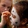 Waikiki MLK parade provides reminder of unity A young girl with dark hair is having her face painted to resemble a Dalmatian dog, featuring white base paint, black spots, a black nose, and whisker lines. An older red-haired child in a green shirt is applying the face paint using a small brush. The scene appears to take place at an outdoor event or fair.