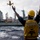 USS Lake Champlain (CG 57) Replenishment-at-Sea A naval personnel wearing a yellow hard hat and a yellow safety vest marked 'CG57' with a 'T' symbol is directing a replenishment-at-sea (RAS) operation with both arms raised. A pallet of supplies is being transferred via a highline/cable system between two naval vessels on open ocean. The operation appears to be a standard underway replenishment (UNREP) between a supply ship and USS Lake Champlain (CG-57).