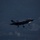 Lightning II strikes Iwakuni, F-35B arrives A military stealth fighter jet, identifiable as an F-35, is captured in low-light/dusk conditions with landing gear deployed, suggesting it is on approach for landing or has just taken off. The aircraft is illuminated by its own navigation and landing lights, including visible green lights and a bright forward landing light. The dark background and motion blur in the lower portion of the image suggest this was taken at twilight or nighttime conditions.