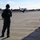 Every clime and place: VMAT-203 Marines maintain expeditionary mindset at MCALF Bogue A military personnel member in flight suit stands on an airfield tarmac observing a fighter jet taxiing in the background. The individual is wearing a backpack and headset, facing away from the camera toward the aircraft. The setting appears to be a military air base surrounded by tree lines under a partly cloudy sky.