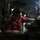 Maintenance A male worker wearing a red uniform and safety glasses is operating a metal lathe or milling machine in a dimly lit industrial workspace. He is leaning in closely to inspect or adjust the machinery, with a bright work light illuminating the work area. The environment appears to be a confined mechanical or engineering space, possibly aboard a naval vessel or industrial facility.